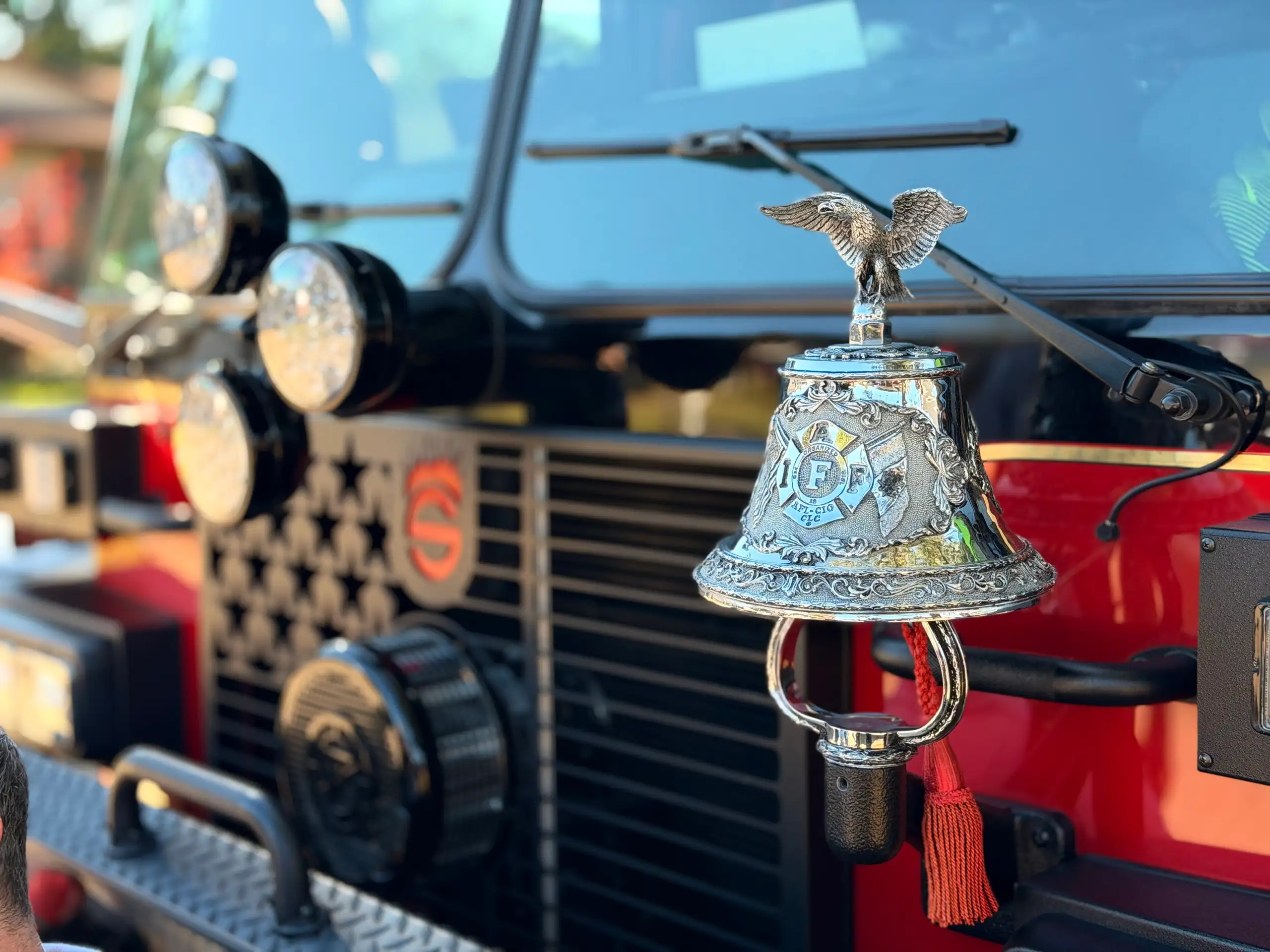 The chrome ceremonial bell of a Maryland Heights engine, topped with an eagle and bearing the IAFF crest, mounted beside the rig's front grille in the sun