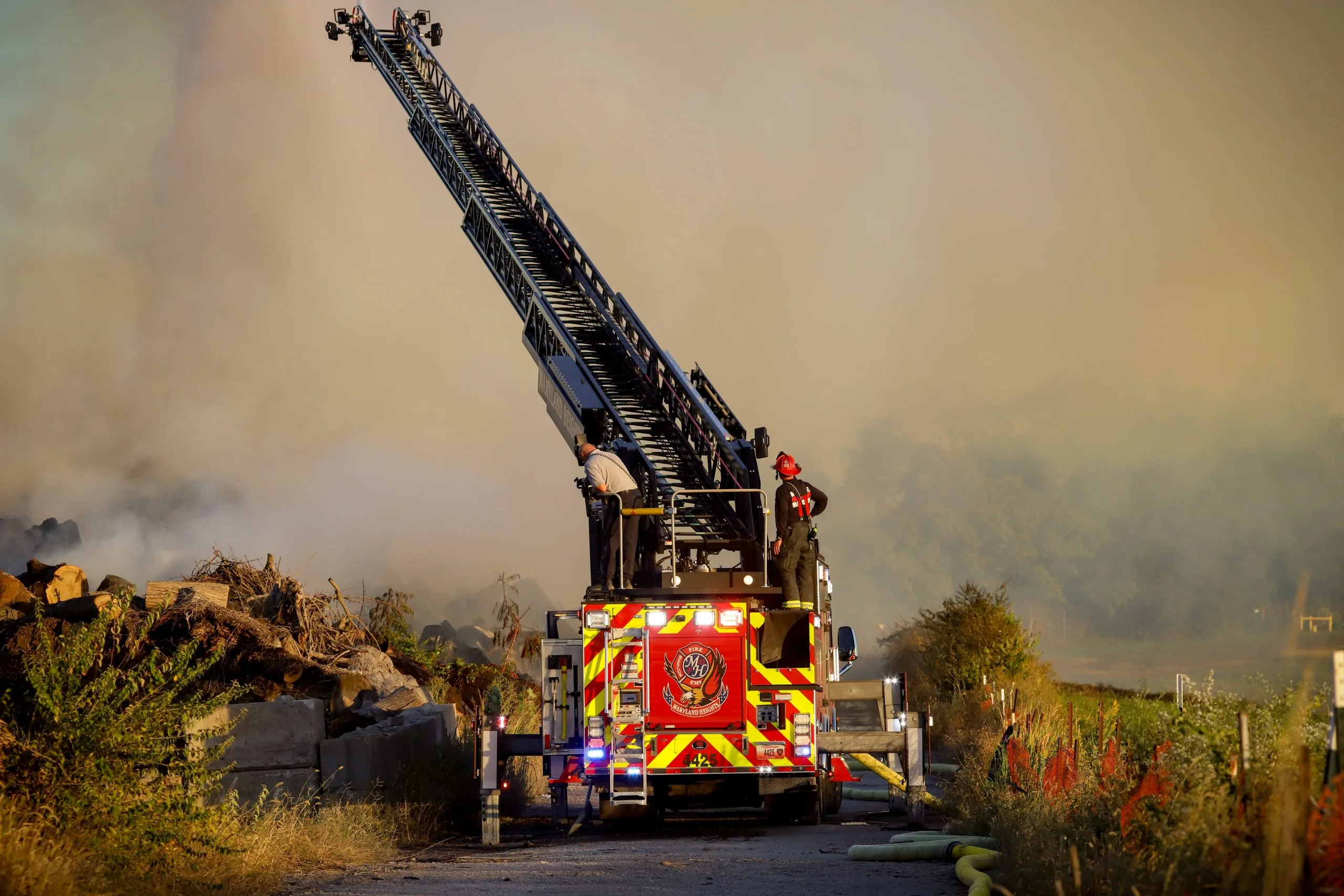 Firefighters atop Rescue Ladder 4425 with its aerial ladder extended into heavy smoke at a fire scene
